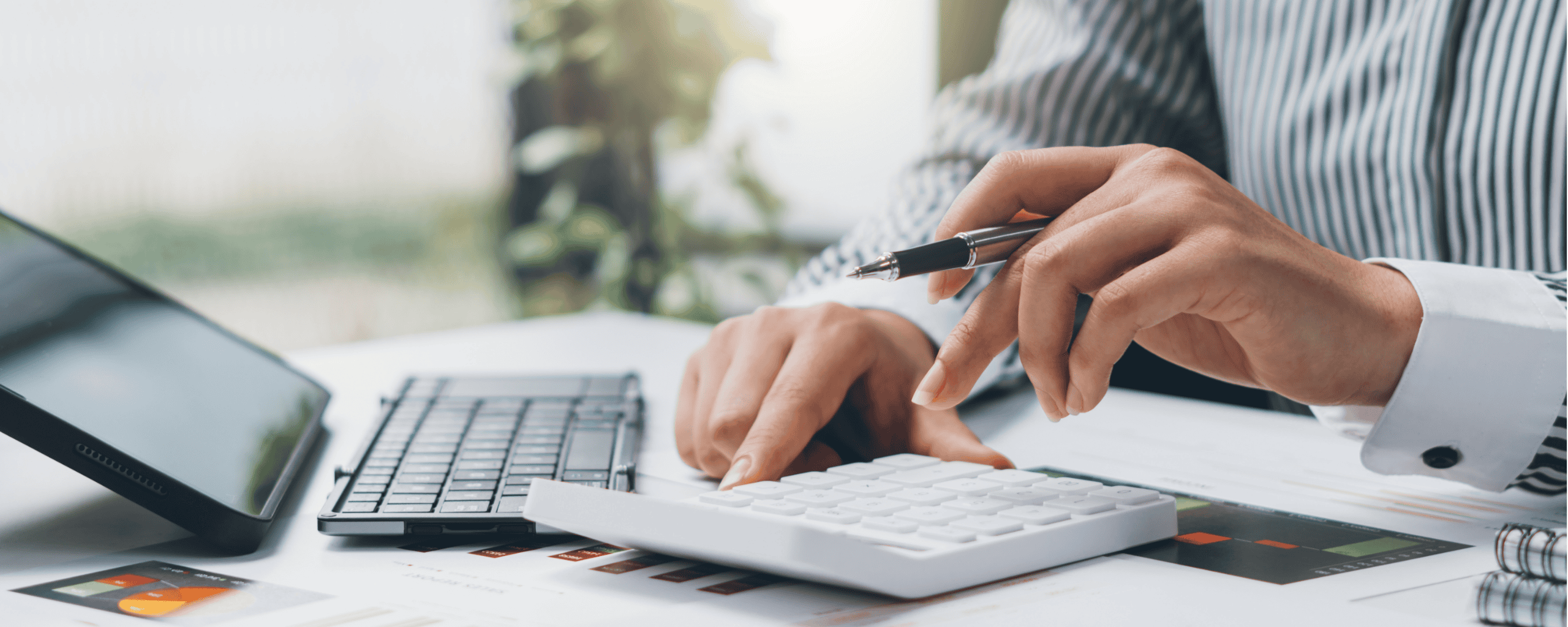 Close-up of a person at a desk using a calculator while holding a pen, with financial documents, charts, a keyboard and a tablet visible. The image represents financial planning, budgeting, accounting or tax calculations in a professional office setting.